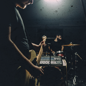 Guy playing electric guitar near camera with mixing board and drummer blurred in the background - grainy image for art